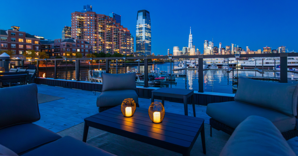 FLOHOM 16 Isaac's Way rooftop lounge with seating and lanterns overlooking Jersey City waterfront and Lower Manhattan skyline
